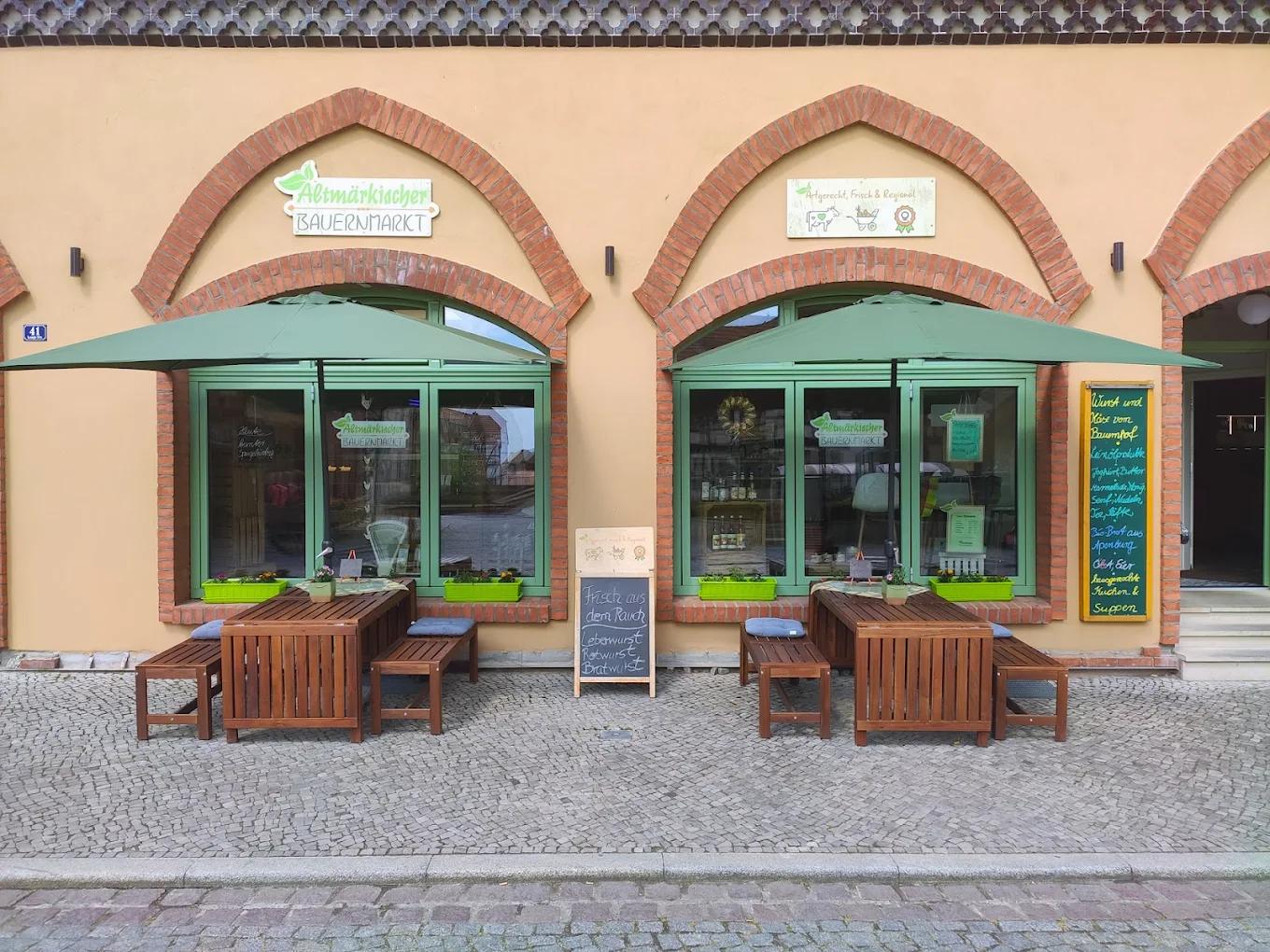 Der Altmärkische Bauernmarkt in der Langen Straße 41 in Tangermünde. Grüne Sonnenschirme vor den Rundbogenfenstern, Sitzbänke auf dem Kopfsteinpflaster, das Ladenschild über dem Fenster.